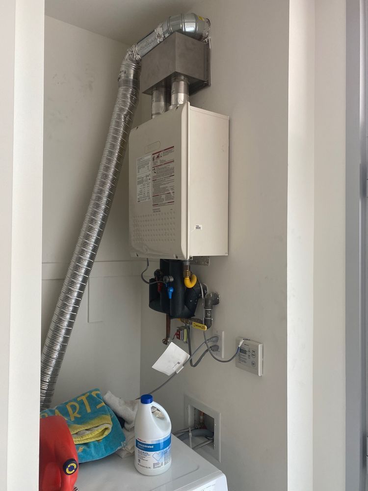 A tankless water heater mounted on a wall in a utility closet with a flexible metal vent pipe and laundry supplies below.