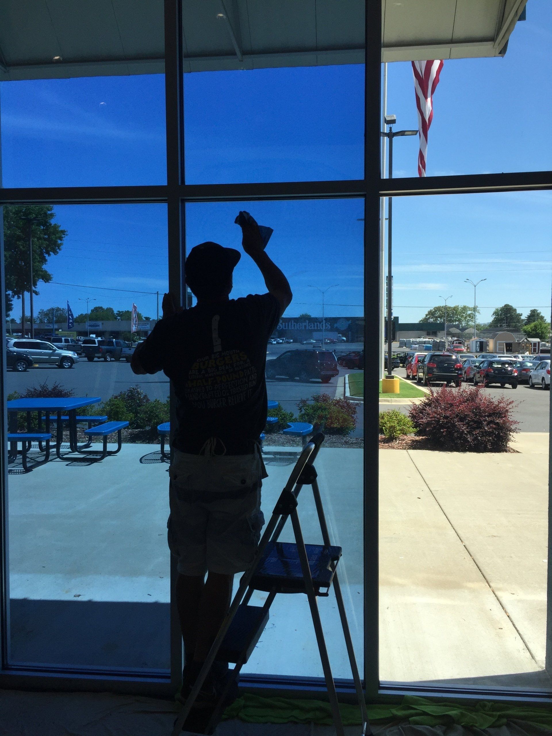 Person applying blue tint to a window; small stepladder in front. A street scene is visible through the untinted portion of the window.