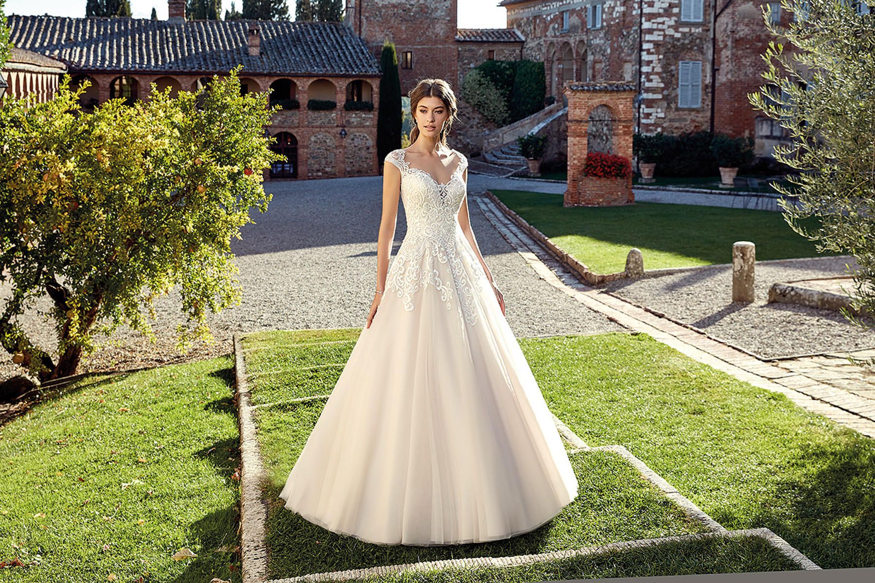 Woman in a wedding dress stands in a garden. She is standing in front of a garden, her dress flowing over the grass.