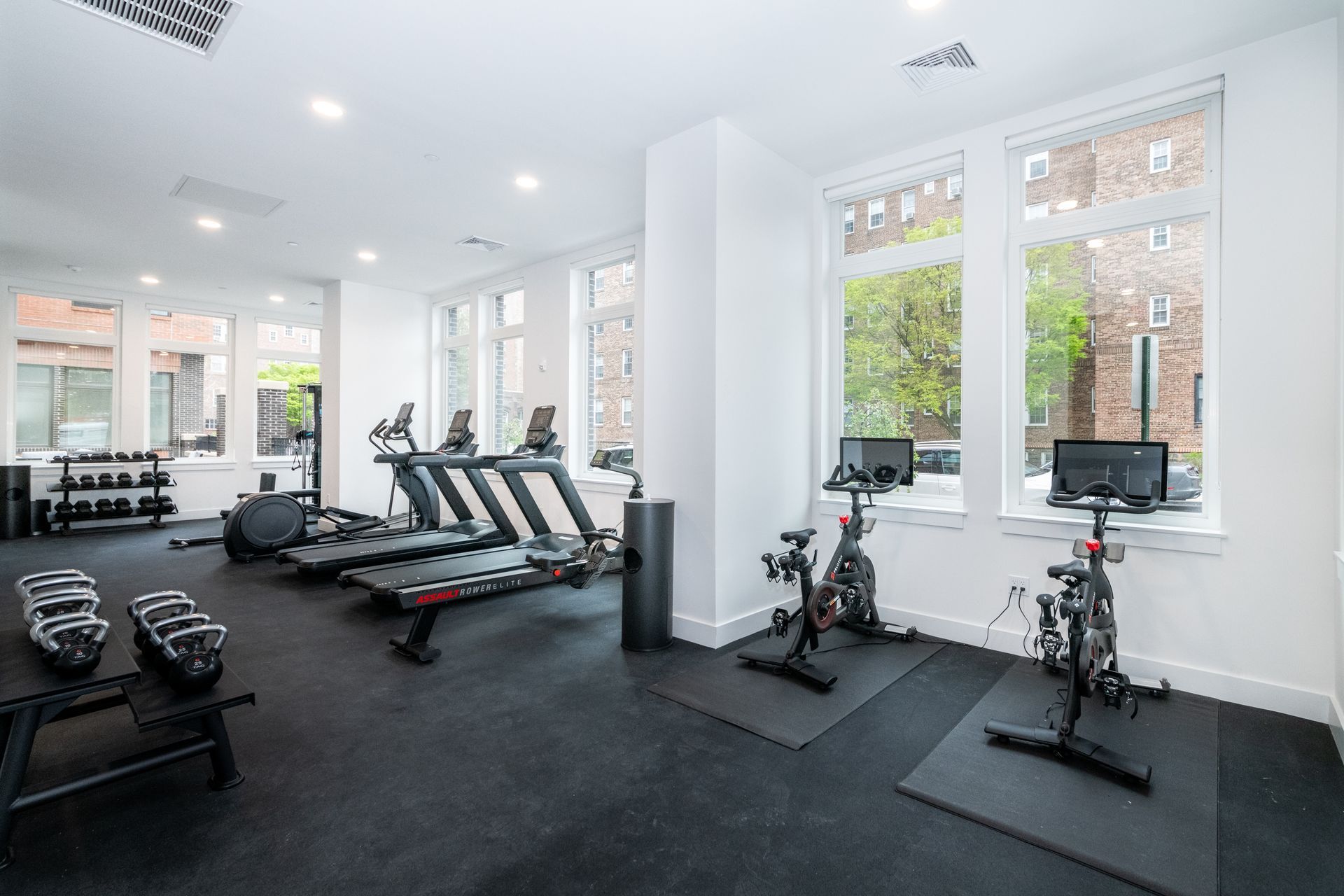 Gym with weights, treadmills, and exercise bikes by large windows. Black flooring and equipment.