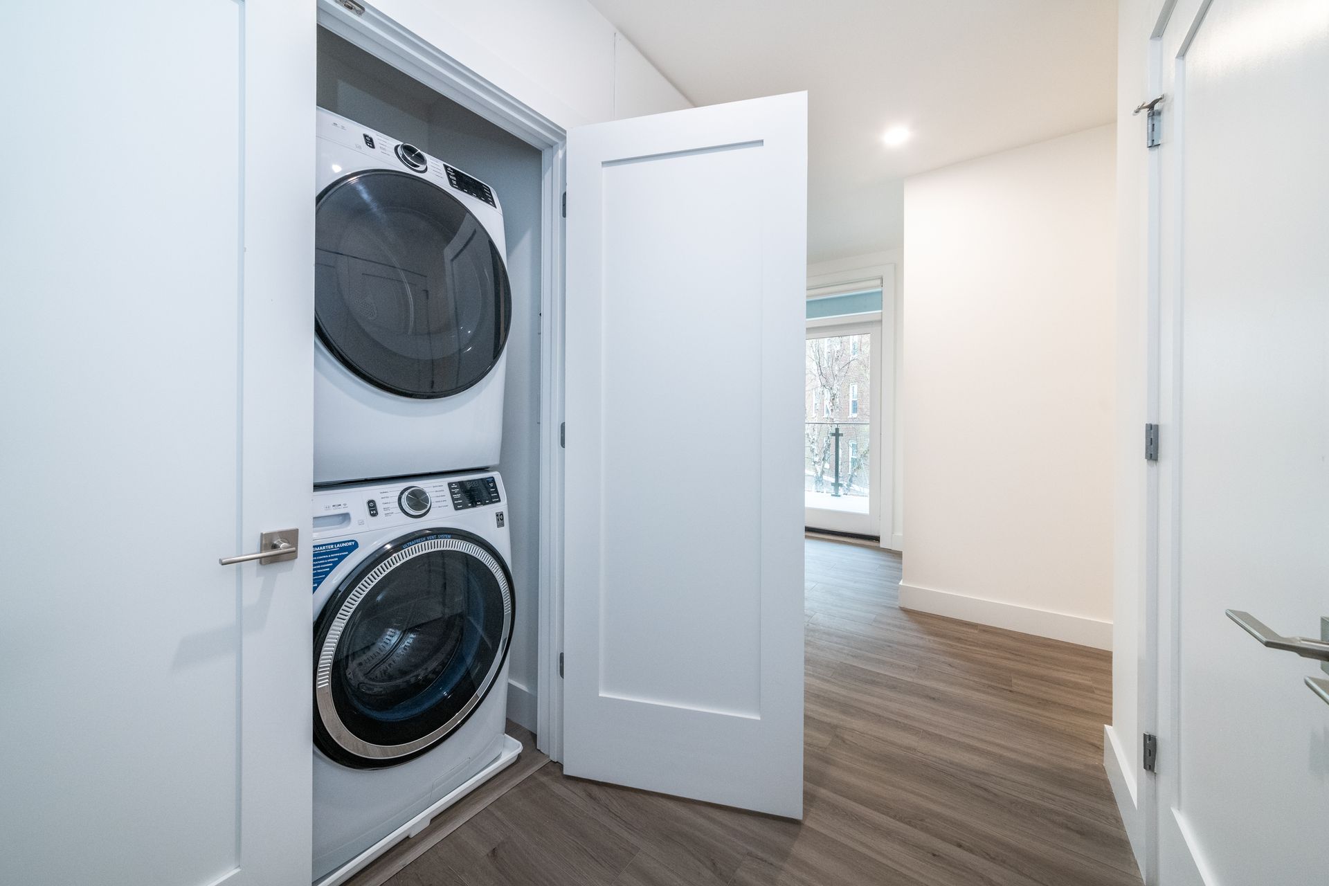 Stacked washer and dryer in a white closet, adjacent to hallway with open door to outdoor scene.
