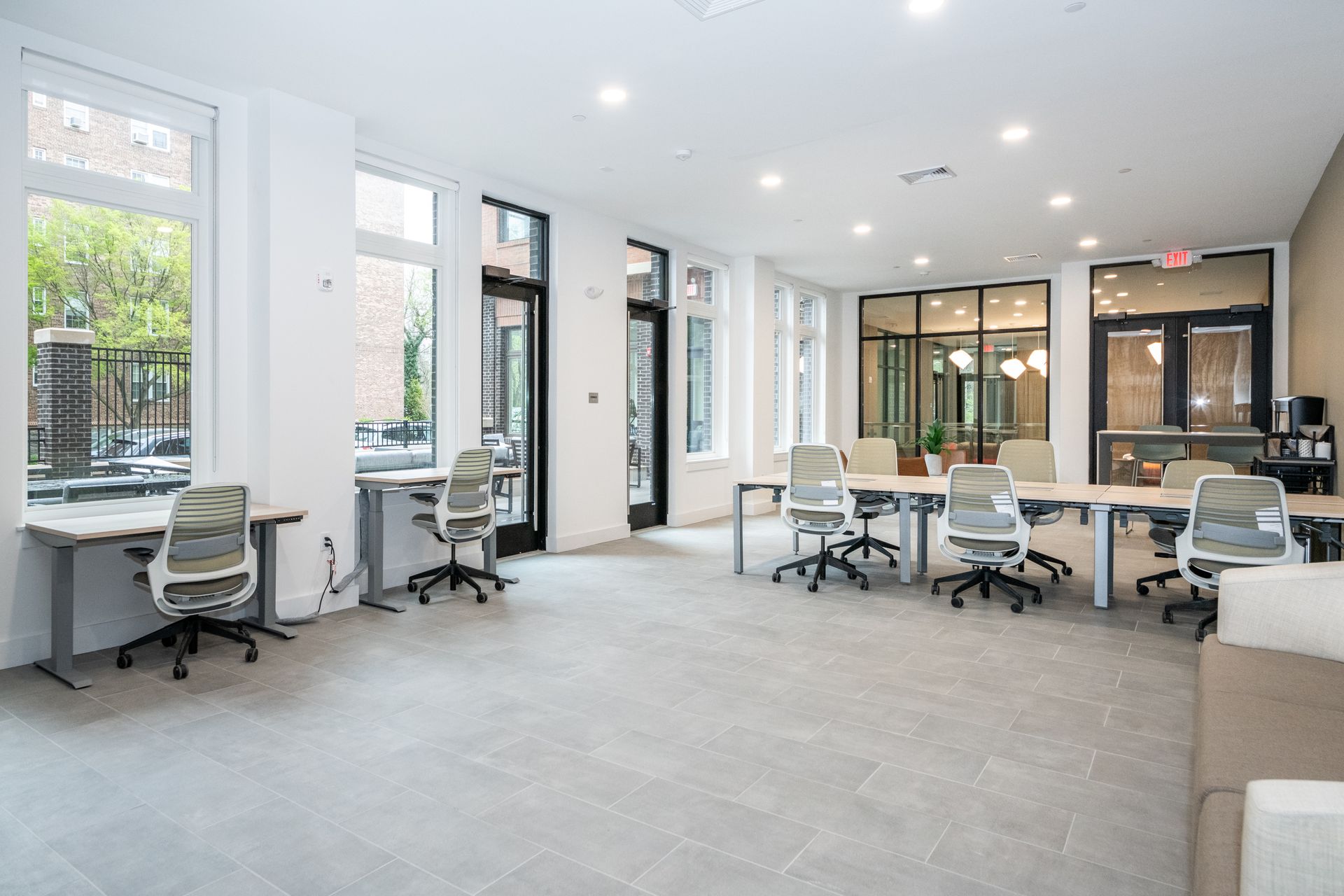 Bright open office with desks, chairs, and windows; a coffee station and outdoor view.
