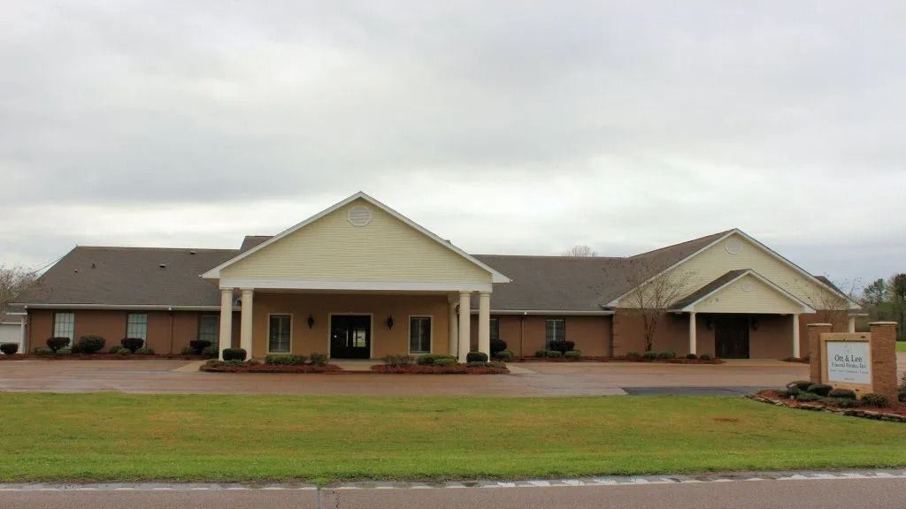Single-story brick building with a central columned entrance, triangular gables, and a sign by the roadside.