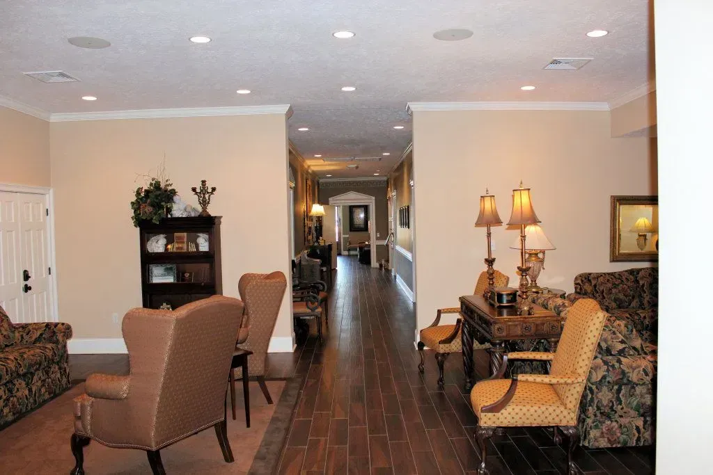 Bright, narrow living room with patterned sofas, chairs, lamps, and a long hallway in the background