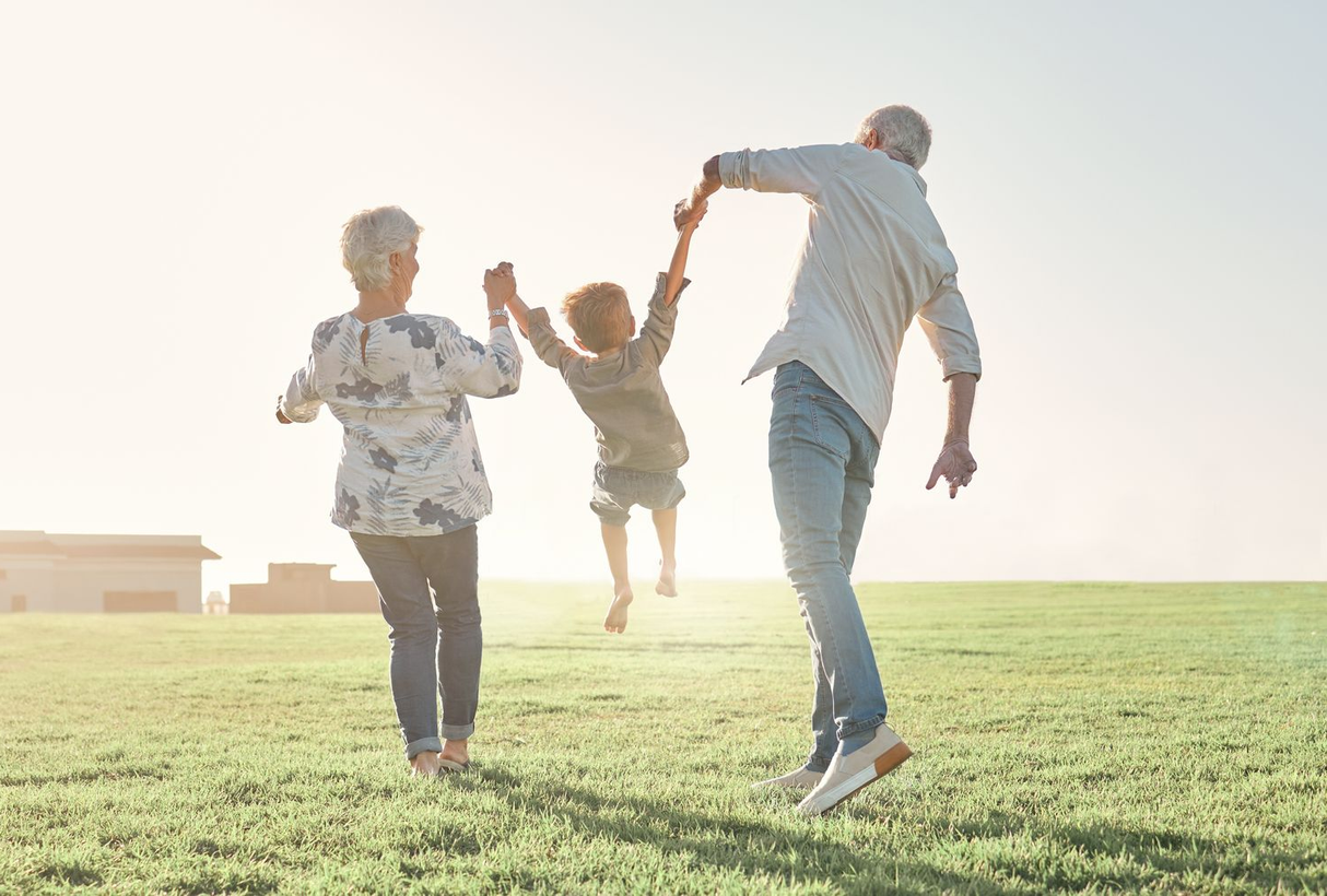 Two adults hold hands with a child and lift them into the air while walking across a sunny grassy field.