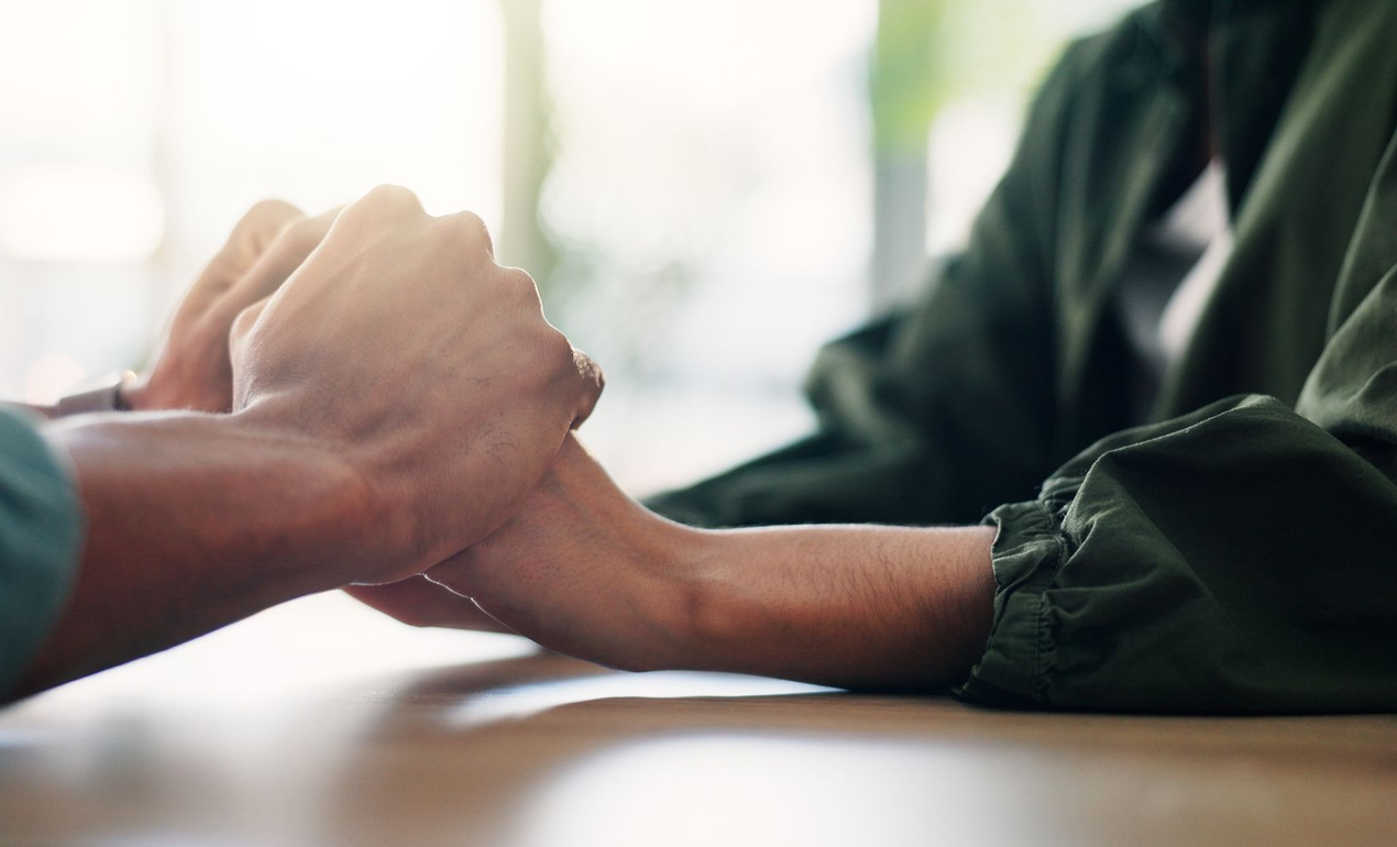 Two people sit at a table, holding hands in a gesture of support and comfort.