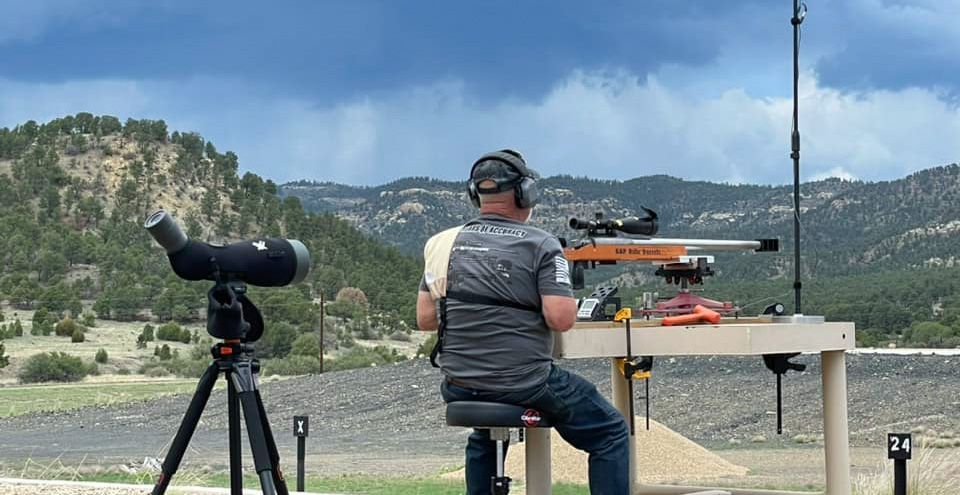 A man using a rifle with a telescope behind him