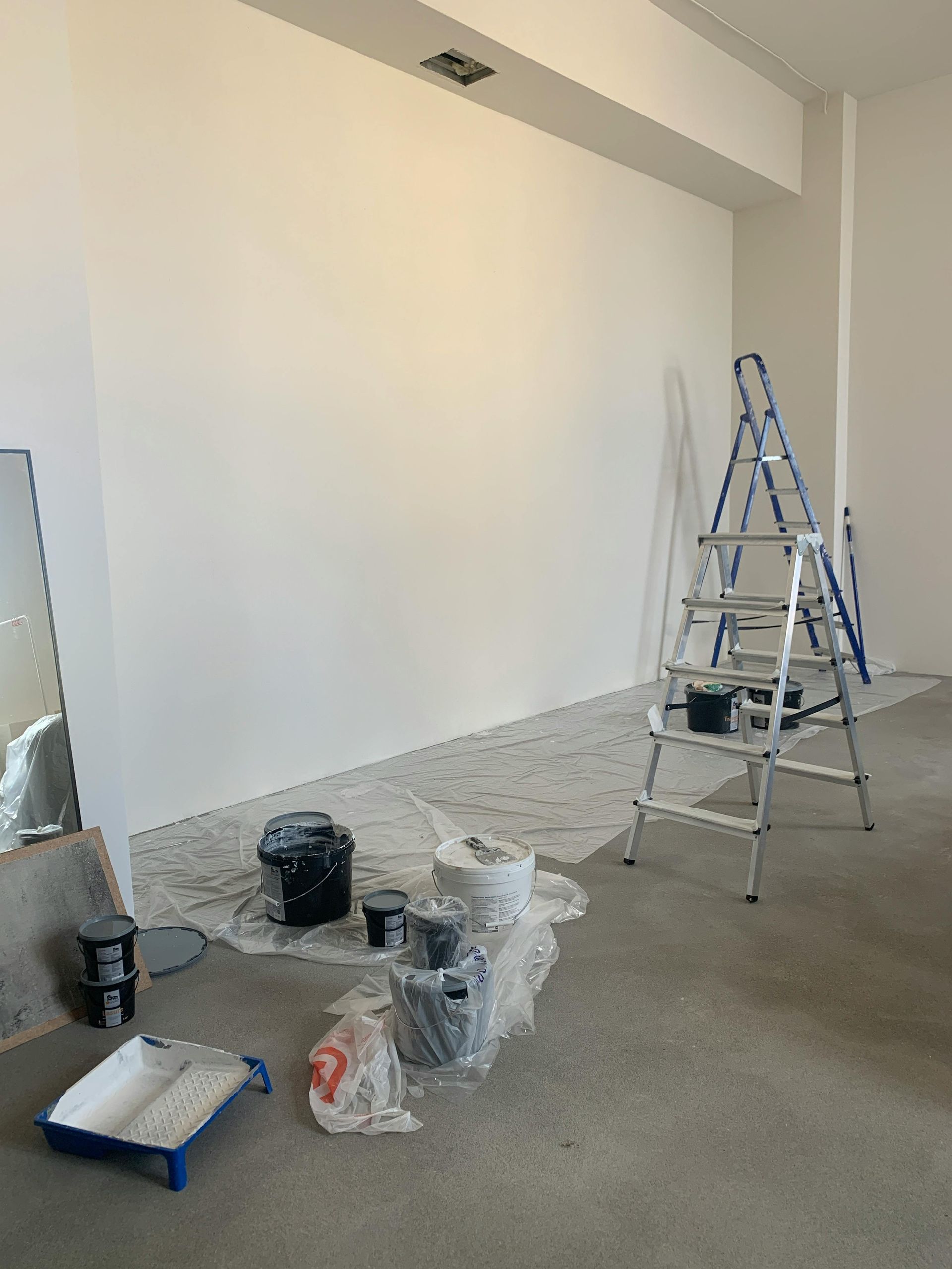 A room with a large white wall, a metal step ladder, paint cans, and a tray on a drop cloth.