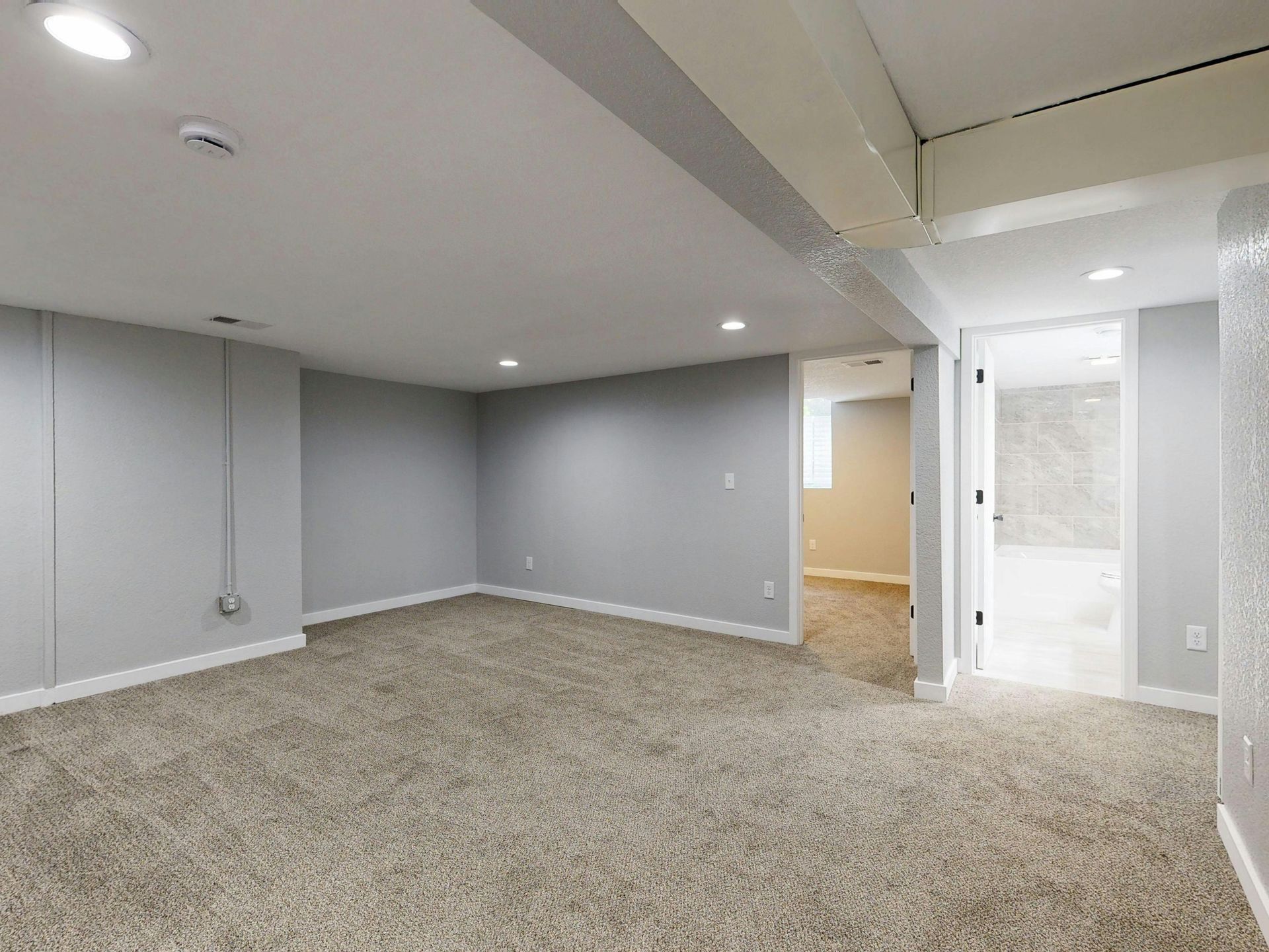 A bright, carpeted basement room with light gray walls and recessed lighting, with an open doorway to a white bathroom.