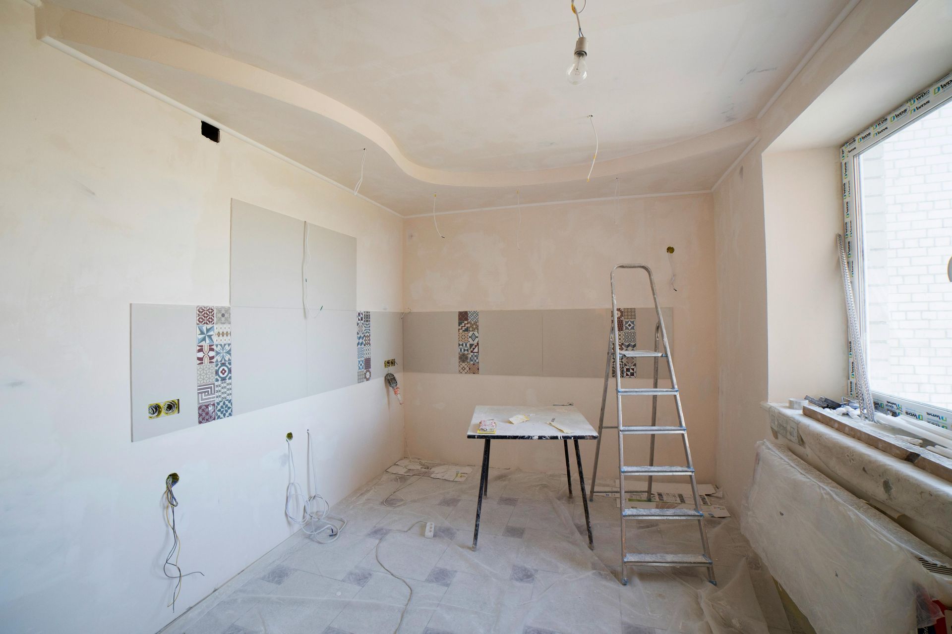 A room under construction with white walls, tiled accents, a step ladder, and a table on a patterned floor.