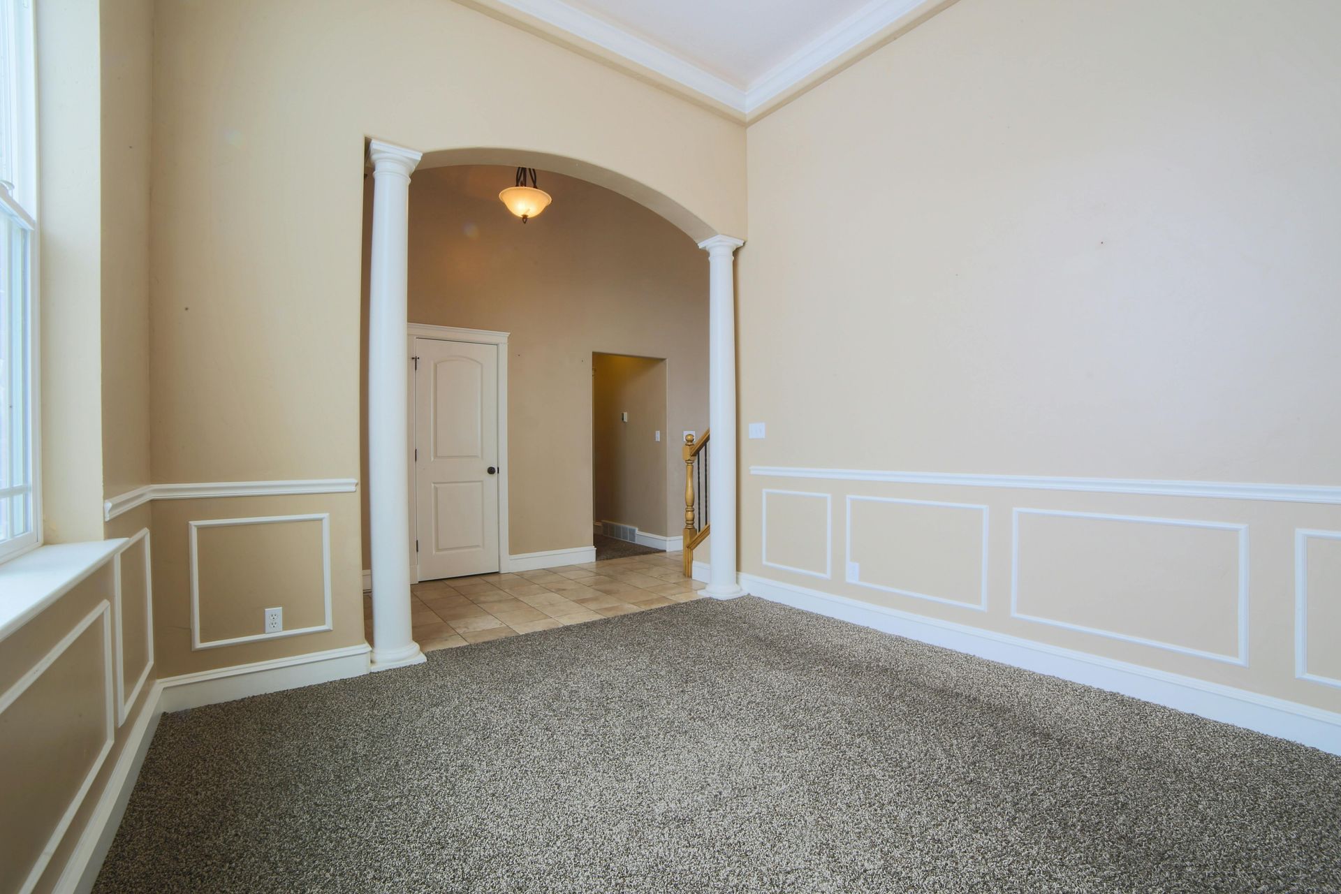 An empty room with beige walls, wainscoting, and carpeted floors, featuring an arched opening with two white columns.
