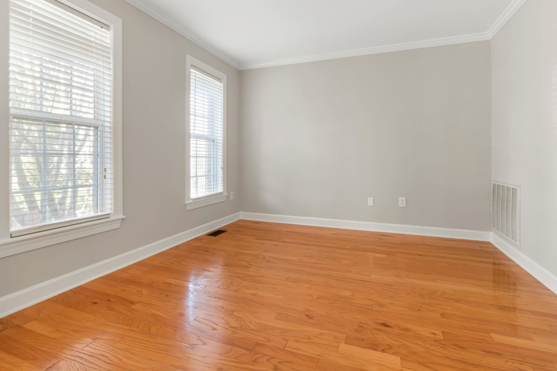 Empty room with light gray walls, hardwood flooring, two windows with white blinds, and white trim.