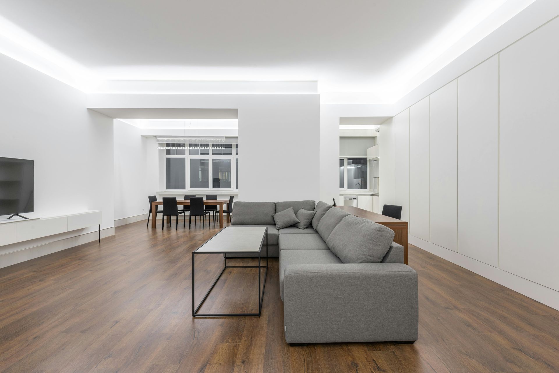 A minimalist, open-concept room with white walls, wood floors, a grey sectional sofa, coffee table, and dining area.