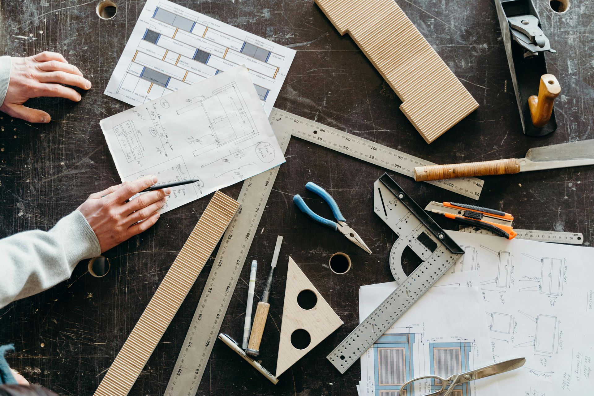A person drafting plans at a dark wooden workbench scattered with tools, rulers, a square, and wood components.