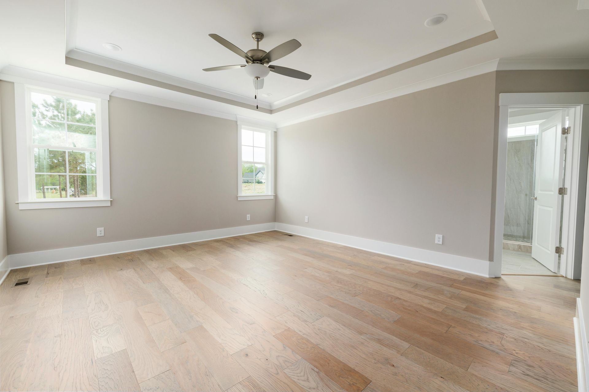 A bright, empty bedroom with light wood floors, gray walls, white trim, a ceiling fan, and an open door to a bathroom.