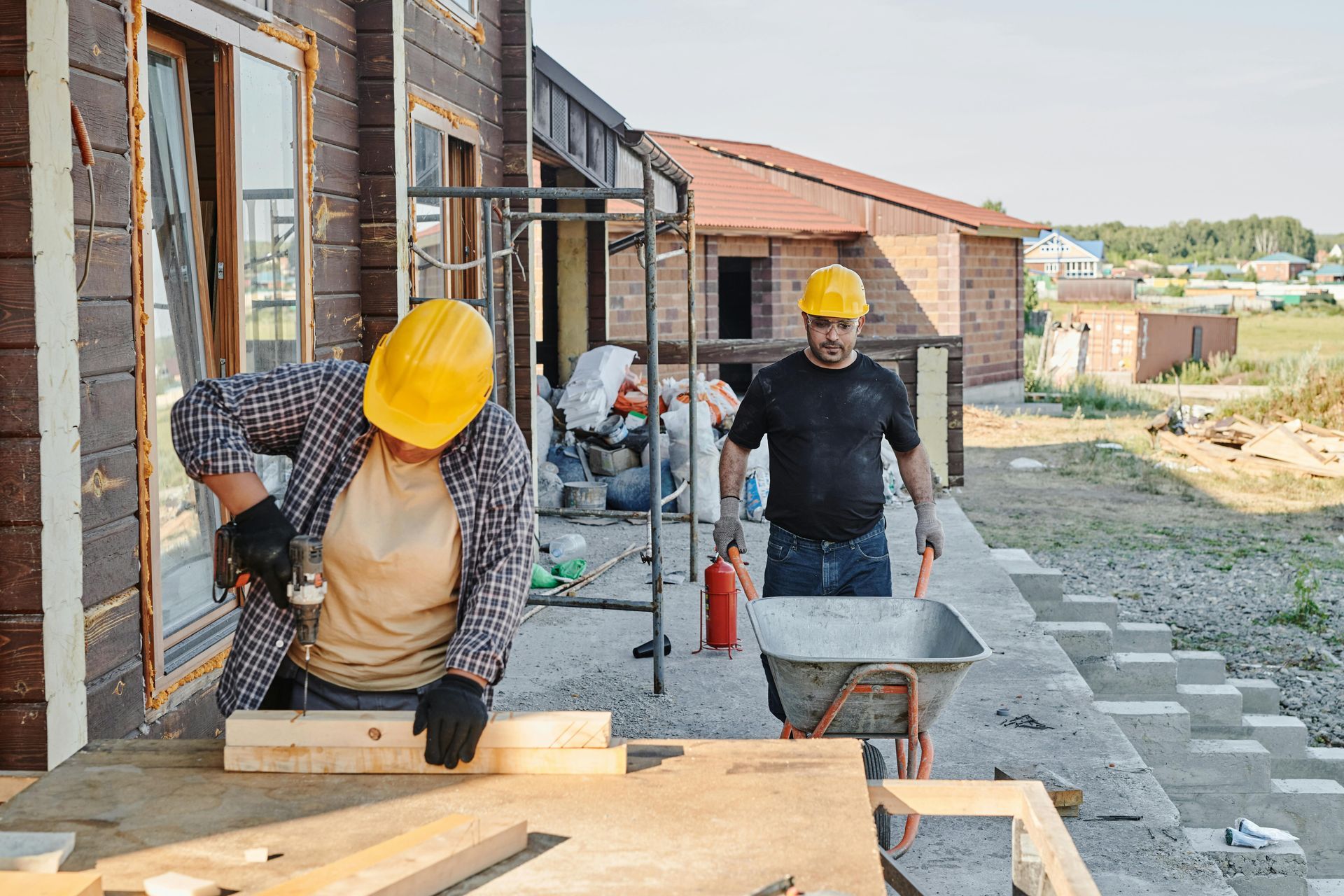 Two construction workers in hard hats work at a house site, one operating a saw on lumber and the other pushing a cart.