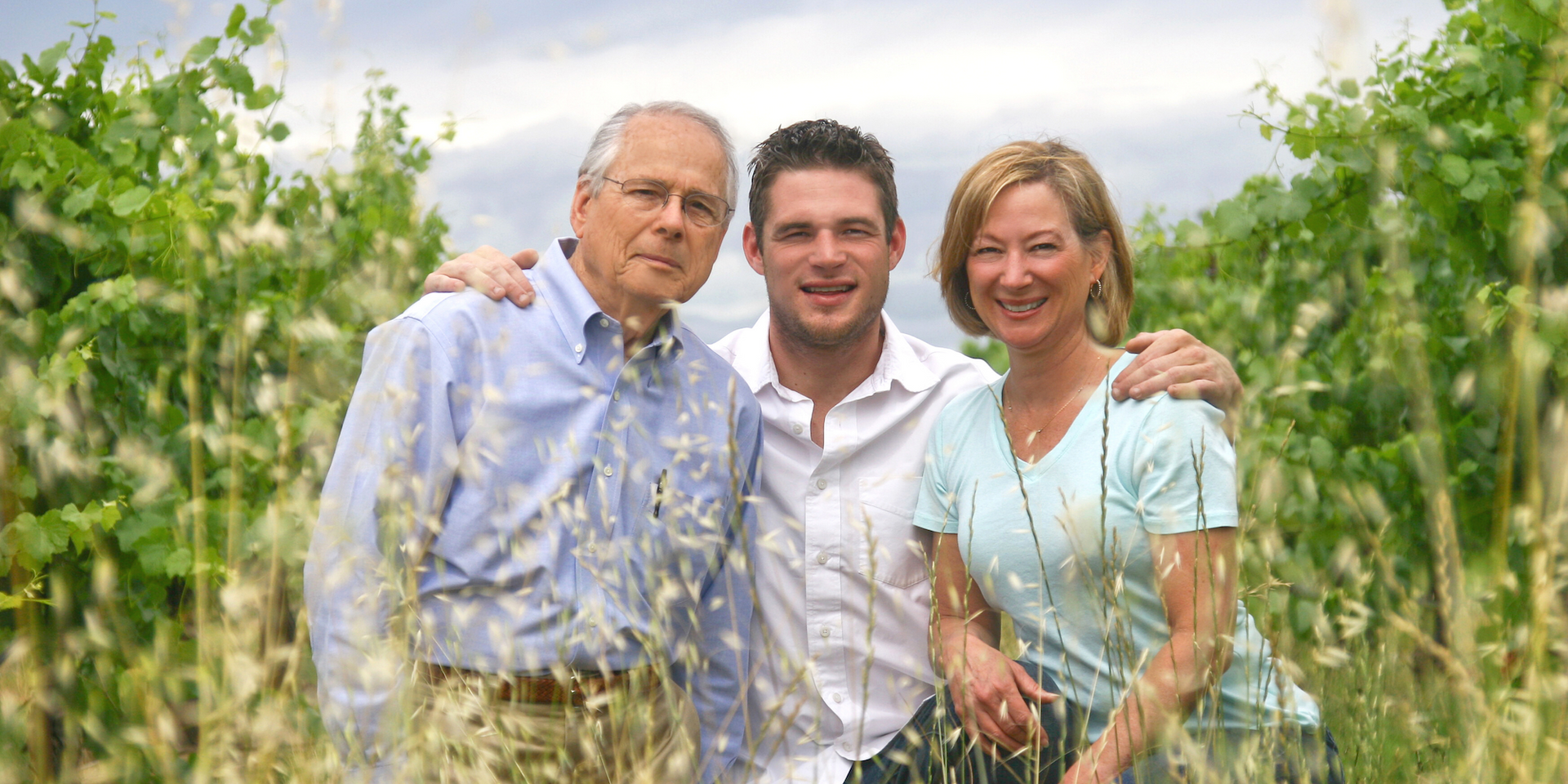 Three people standing together in a vineyard, smiling and posing for a photo outdoors.