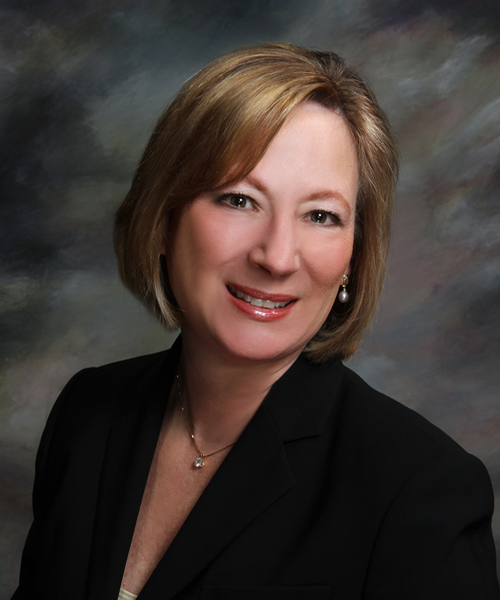 A headshot of a person with shoulder-length blonde hair, smiling, wearing a black blazer and a delicate necklace.