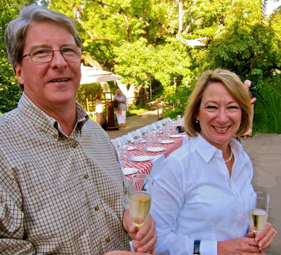 A man and woman holding glasses of champagne at an outdoor table setting with green trees in the background.