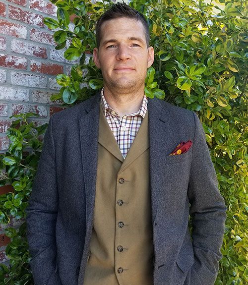 A person wears a dark gray blazer, tan vest, and patterned shirt, standing in front of a brick wall and green foliage.