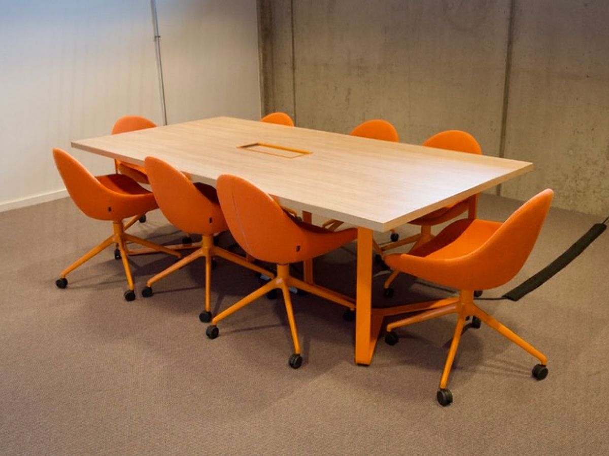 Ein rechteckiger Konferenztisch aus hellem Holz mit acht orangefarbenen, drehbaren Bürostühlen in einem Raum mit Betonwänden.