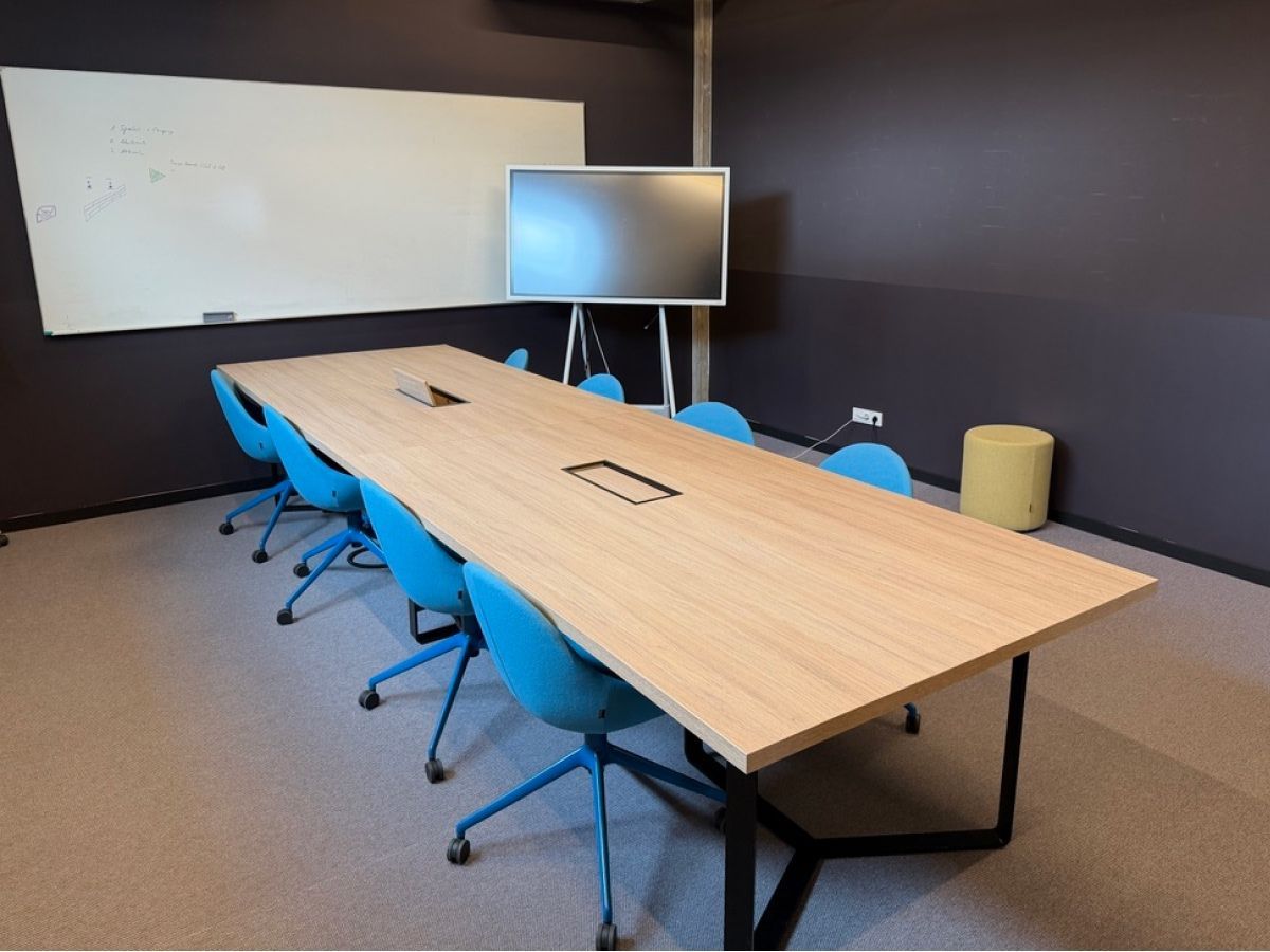 Ein moderner Besprechungsraum mit einem langen hellen Holztisch, umgeben von sechs blauen Stühlen, einer Whiteboard-Tafel und einem Bildschirm.
