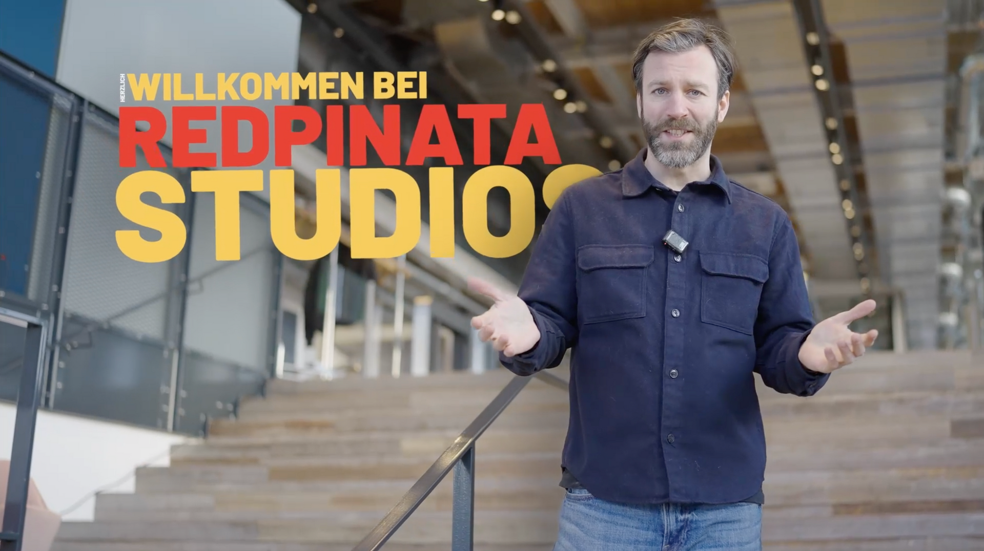 Ein Mann steht auf einer Treppe in einem hellen, modernen Studio und begrüßt die Zuschauer mit einer Geste bei Redpinata Studios.