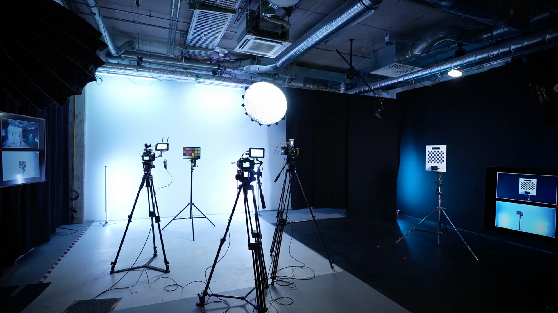 Ein Studio-Set mit mehreren Kameras auf Stativen, einem großen weißen Hintergrund, Beleuchtungsequipment und Monitoren an schwarzen Wänden.