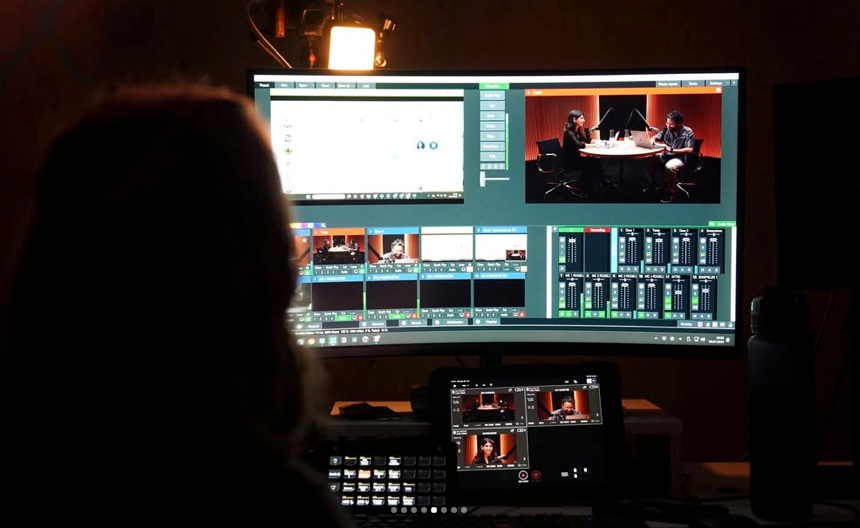Eine Person sitzt in einem dunklen Raum vor einem großen Computermonitor, auf dem Live-Videobearbeitungssoftware und Podcast-Aufnahmen angezeigt werden.
