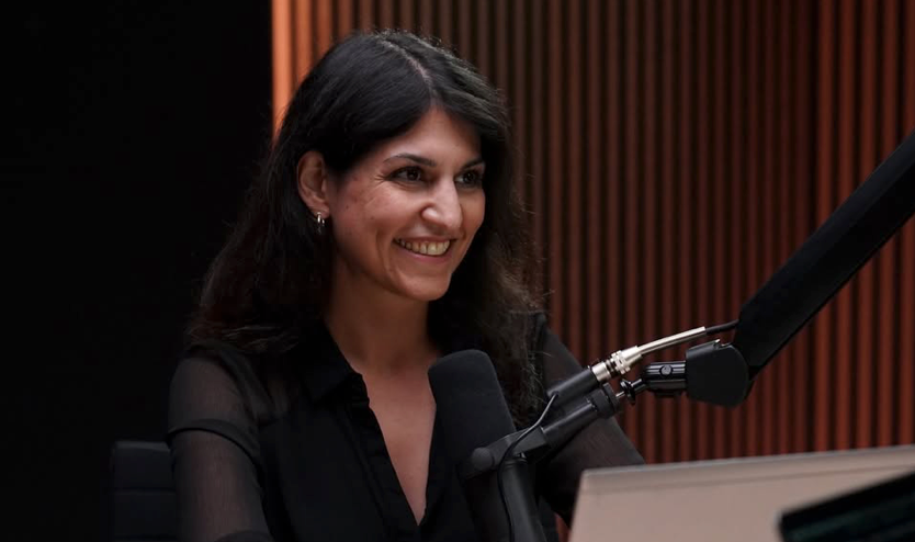 Eine Person lächelt, während sie in einem Studio vor vertikalen, holzgetäfelten Wänden in ein Mikrofon spricht.