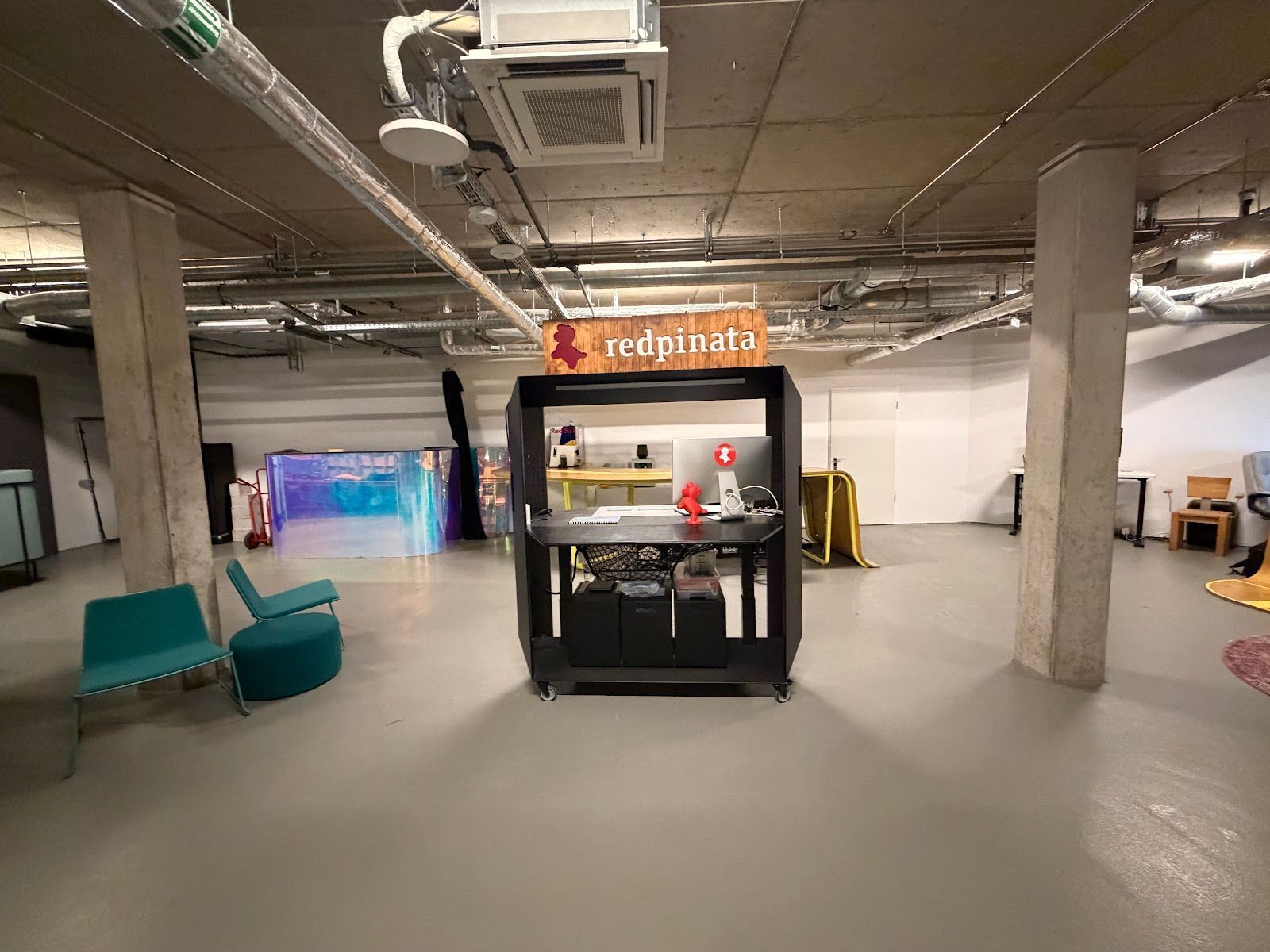 Ein modernes, im Industriestil gehaltenes Kellerstudio mit offenem Grundriss, einem zentralen dunklen Schreibtisch und türkisfarbenen Sitzmöbeln.