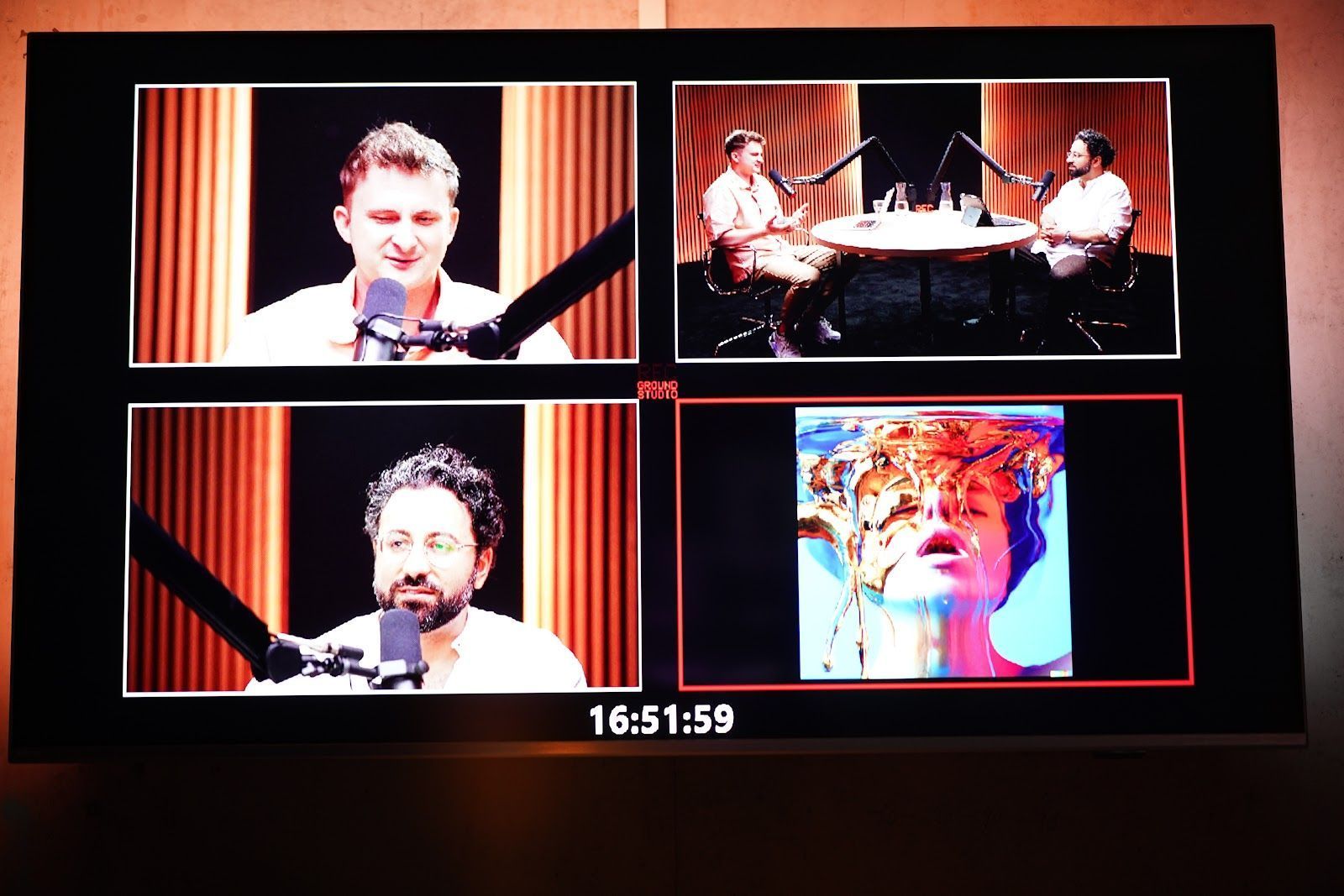 Auf einem Monitor werden vier Videobilder angezeigt: drei zeigen zwei Männer, die in Mikrofone sprechen, und eines zeigt eine farbenfrohe Nahaufnahme.