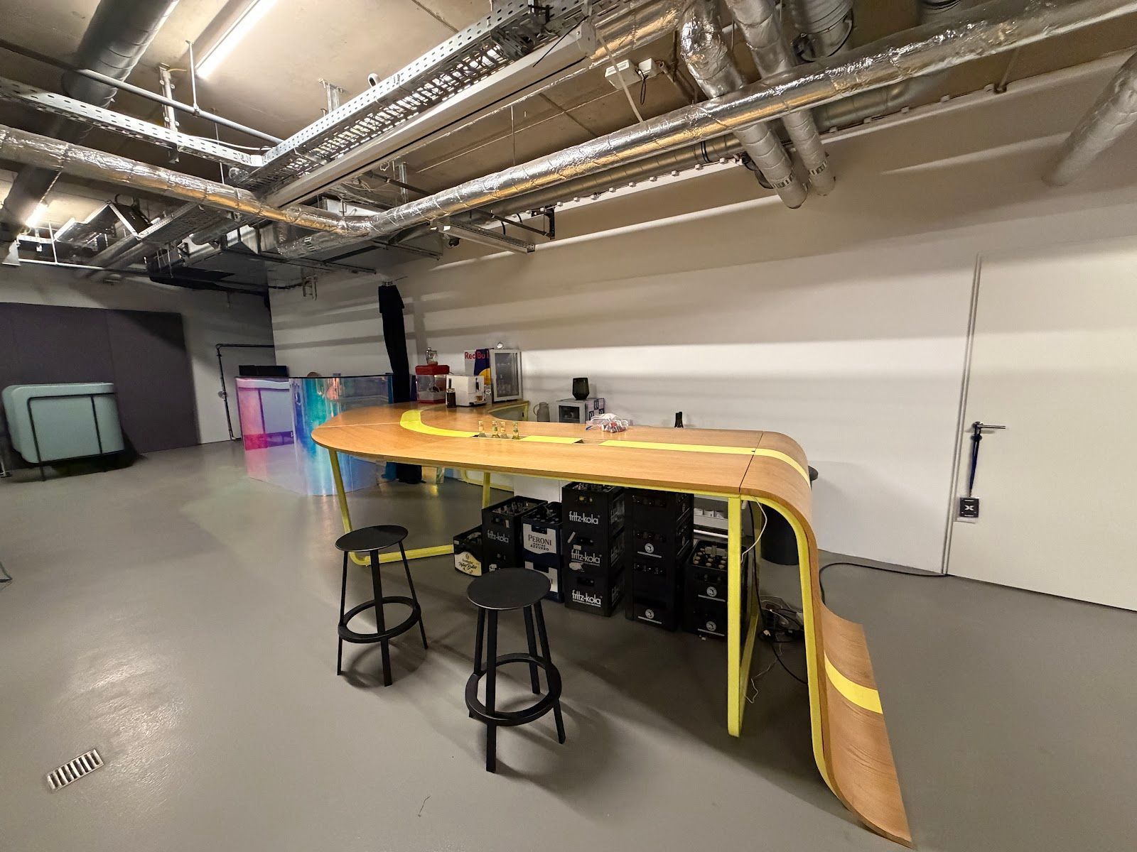 In einem Raum mit freiliegenden Industriedeckenrohren und zwei schwarzen Barhockern steht ein gelber, geschwungener Holzarbeitstisch.