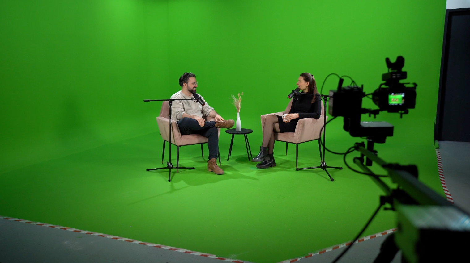 Zwei Personen sitzen auf Stühlen in einem Greenscreen-Studio und unterhalten sich mit Mikrofonen.