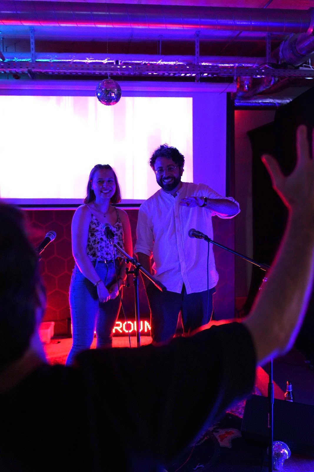 Zwei Personen stehen auf einer Bühne vor einer hell erleuchteten Leinwand; im Vordergrund ist der erhobene Arm einer Person zu sehen.