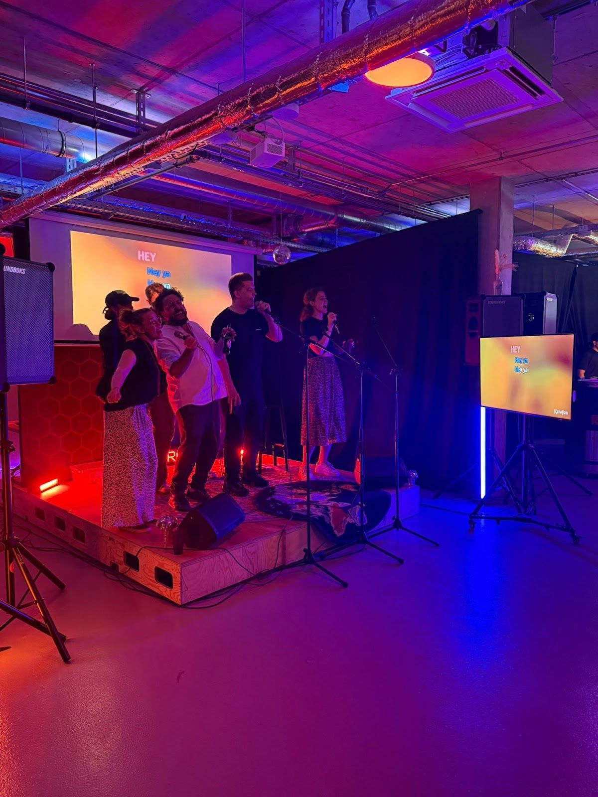 Vier Personen treten auf einer kleinen, hell erleuchteten Bühne in einem schwach beleuchteten Veranstaltungsort im Industriestil mit bunter Neonbeleuchtung auf.