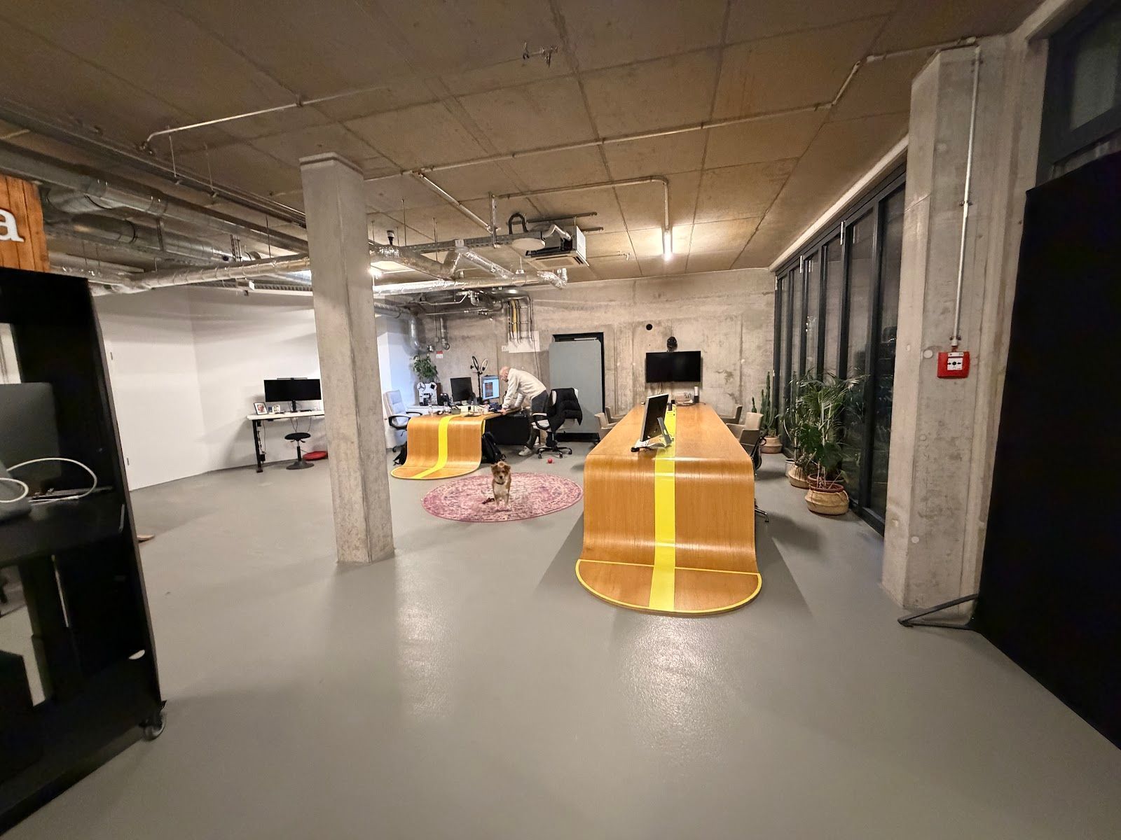 Modernes Industriebüro mit Betonpfeilern, offener Decke, gelb gestreiften Holztischen und großen Fenstern.