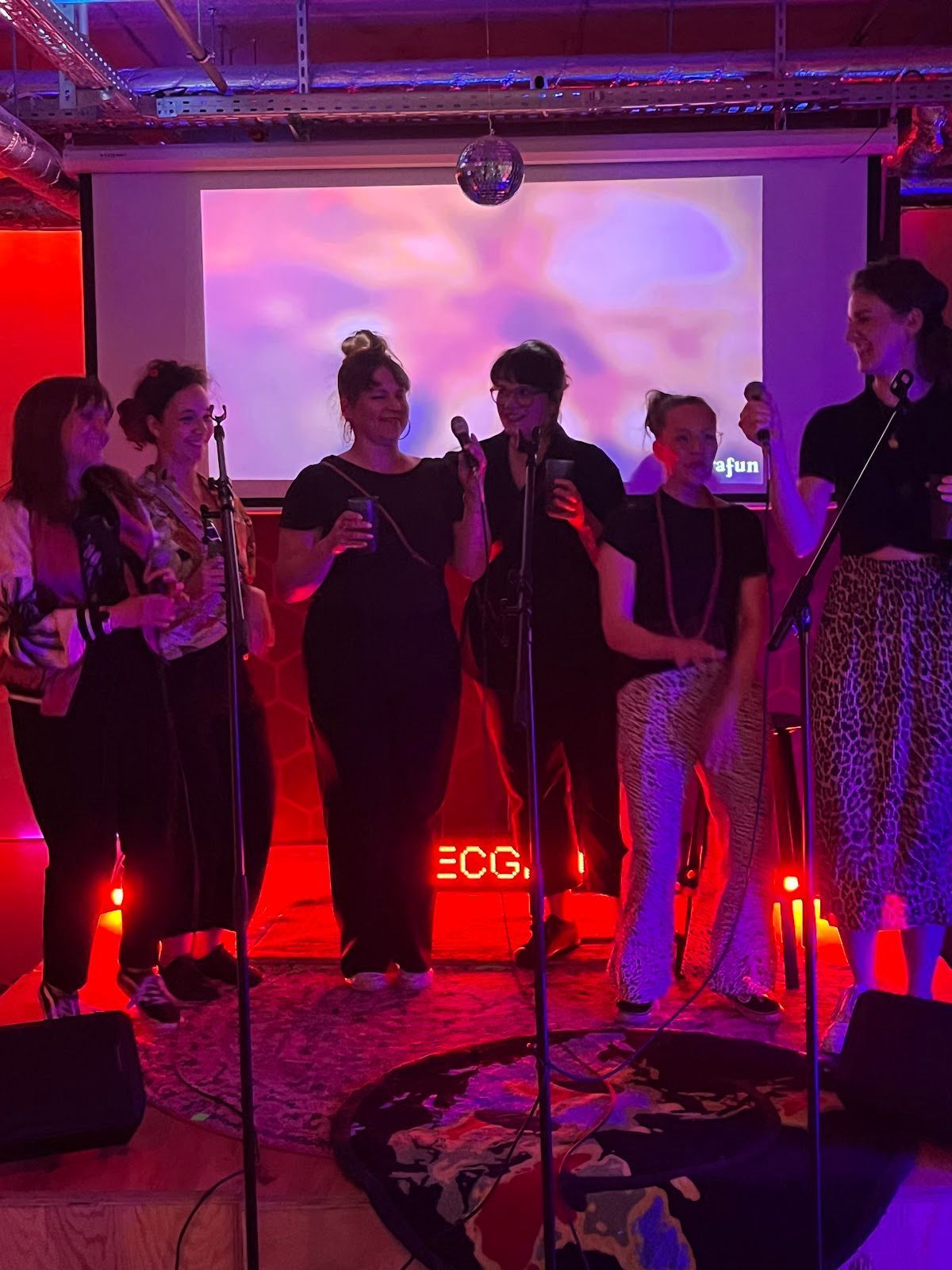 Sechs Personen treten auf einer Bühne auf, im Hintergrund befindet sich eine hell erleuchtete Leinwand, die in rotes und violettes Bühnenlicht getaucht ist.