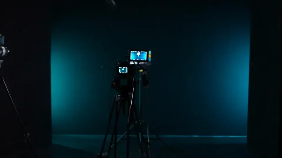 Eine professionelle Videokamera auf einem Stativ ist auf einen schwach beleuchteten Studiohintergrund gerichtet, der von weichem, bläulichem Licht erhellt wird.