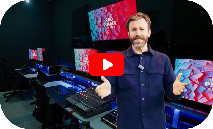 Eine Person gestikuliert in einem Studio mit mehreren Monitoren, auf denen farbenfrohe Grafiken und ein zentraler roter YouTube-Wiedergabeknopf angezeigt werden.