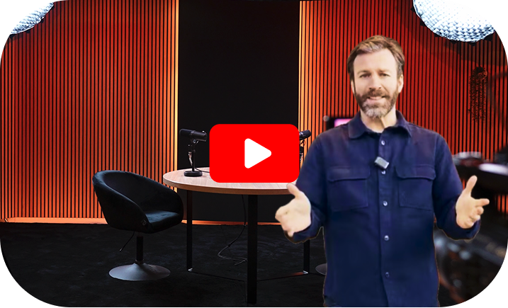 Ein Mann mit Bart, der ein blaues Hemd trägt, steht gestikulierend in einem Studio mit orangefarbenen vertikalen Lamellen und einem Play-Button-Overlay.