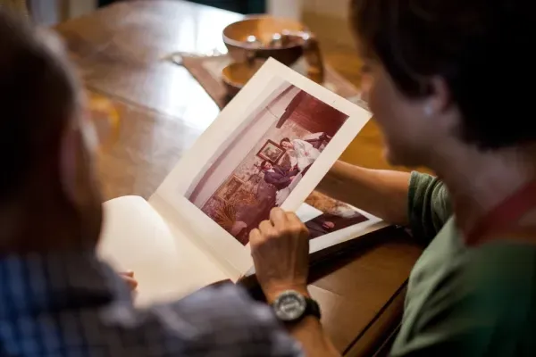 Two people look at a photograph inside a book while sitting at a wooden table.