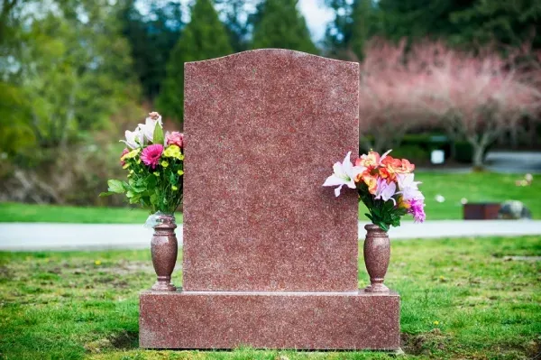 A blank red granite headstone stands in a cemetery with a flower bouquet on each side against a blurred green background.