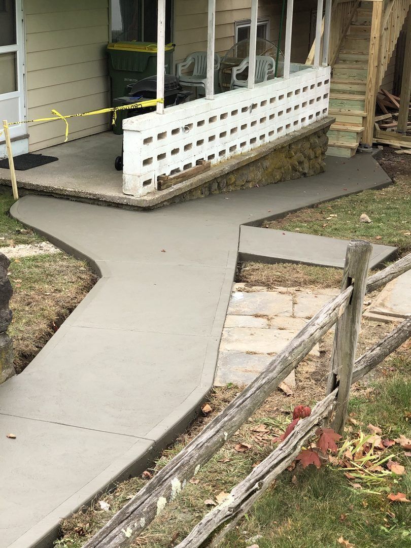 A concrete walkway leading to a porch of a house next to a wooden fence.