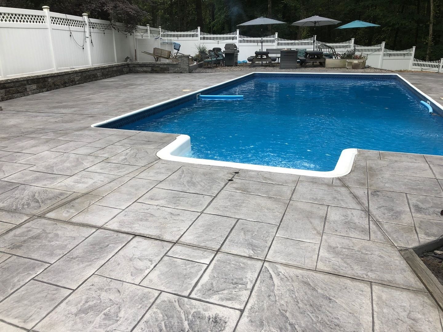 A large swimming pool is surrounded by a concrete patio and a white fence.