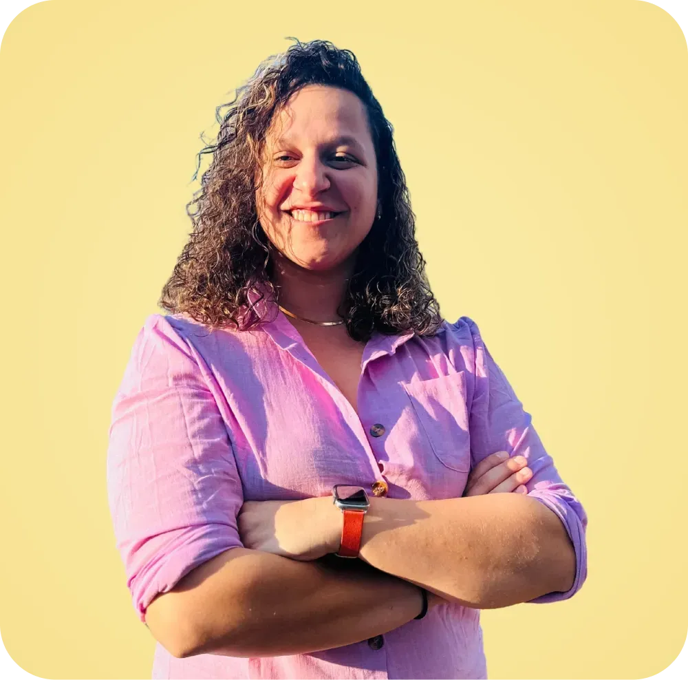 A woman in a pink shirt is standing with her arms crossed and smiling.