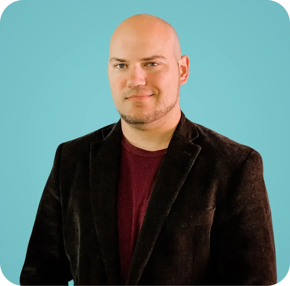A bald man wearing a brown jacket and a red shirt is standing in front of a blue background.