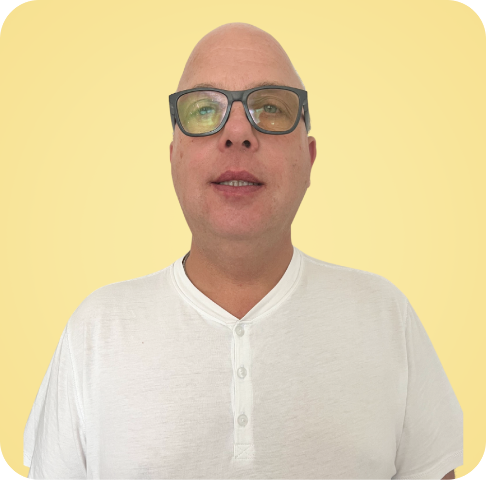 A bald man wearing glasses and a white shirt is standing in front of a yellow background.