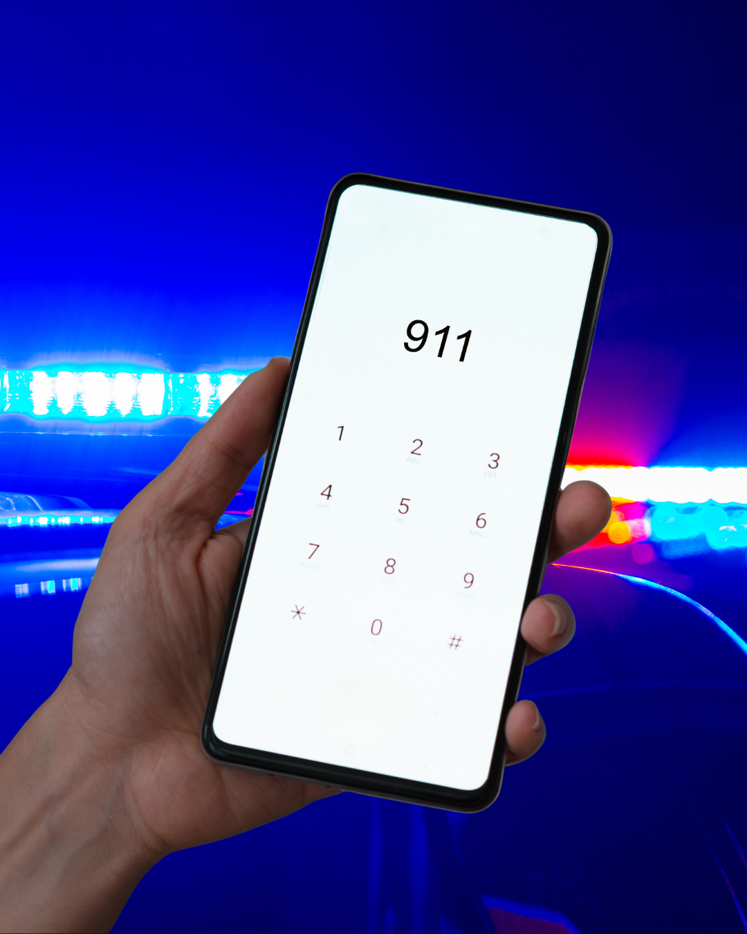 Police car red and blue sirens in the background with a hand holding a phone that shows 911 being called.
