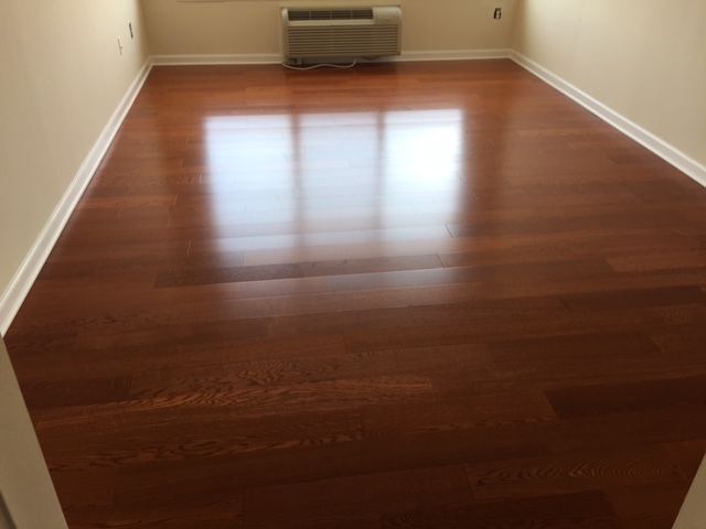 Shiny, reddish-brown hardwood floor in a room with a window reflecting light, beige walls, and white trim.